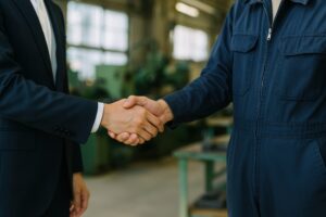 工場内で濃紺の作業服の作業員と握手するビジネスマン