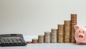 白壁背景に並ぶ多量の硬貨と貯金箱と電卓