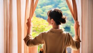窓辺でカーテンを開ける女性が外を眺めるシーン