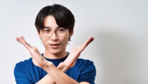 白背景で青いTシャツとメガネをかけて腕をクロスしてNGサインを示す人
