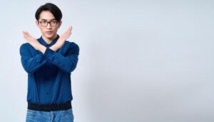 白背景で青いシャツとメガネをかけて腕をクロスしてNGサインを示す人