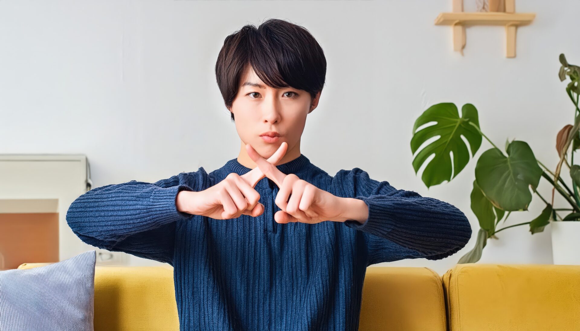 室内で青いトップスを着て指でバツ印を作りNGサインを示す人