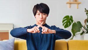 室内で青いトップスを着て指でバツ印を作りNGサインを示す人