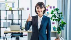 観葉植物やホワイトボードがあるオフィスで親指を立てる笑顔の若い女性