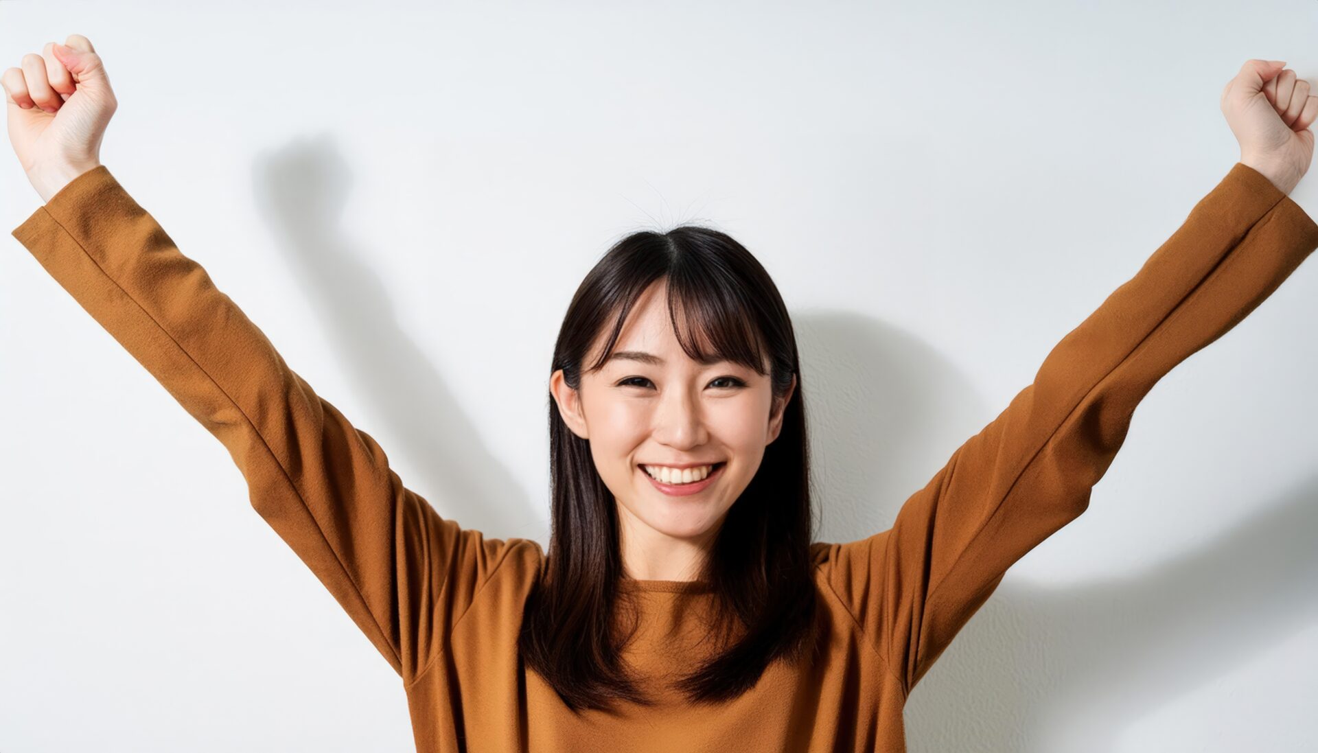 白背景で拳を上げる笑顔の若い女性（ブラウンのトップス）