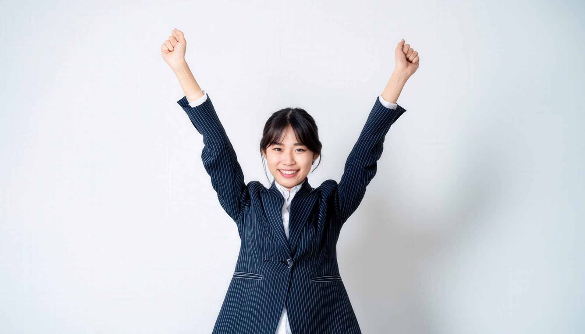白背景で腕を高く上げる笑顔の若い女性（ストライプのスーツ）
