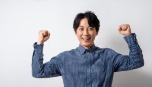 白背景でガッツポーズをする笑顔の若い男性（ストライプシャツ・前髪あり）