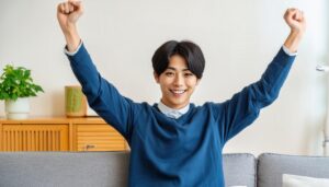 室内でガッツポーズをする若い男性（ネイビートップス）