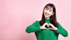 ハートマークを作る若い女性（グリーントップス・ピンク背景）