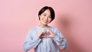 ピンク背景でハートマークを作る若い女性（ストライプシャツ）