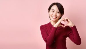 ピンク背景でハートマークを作る若い女性（レッドトップス）