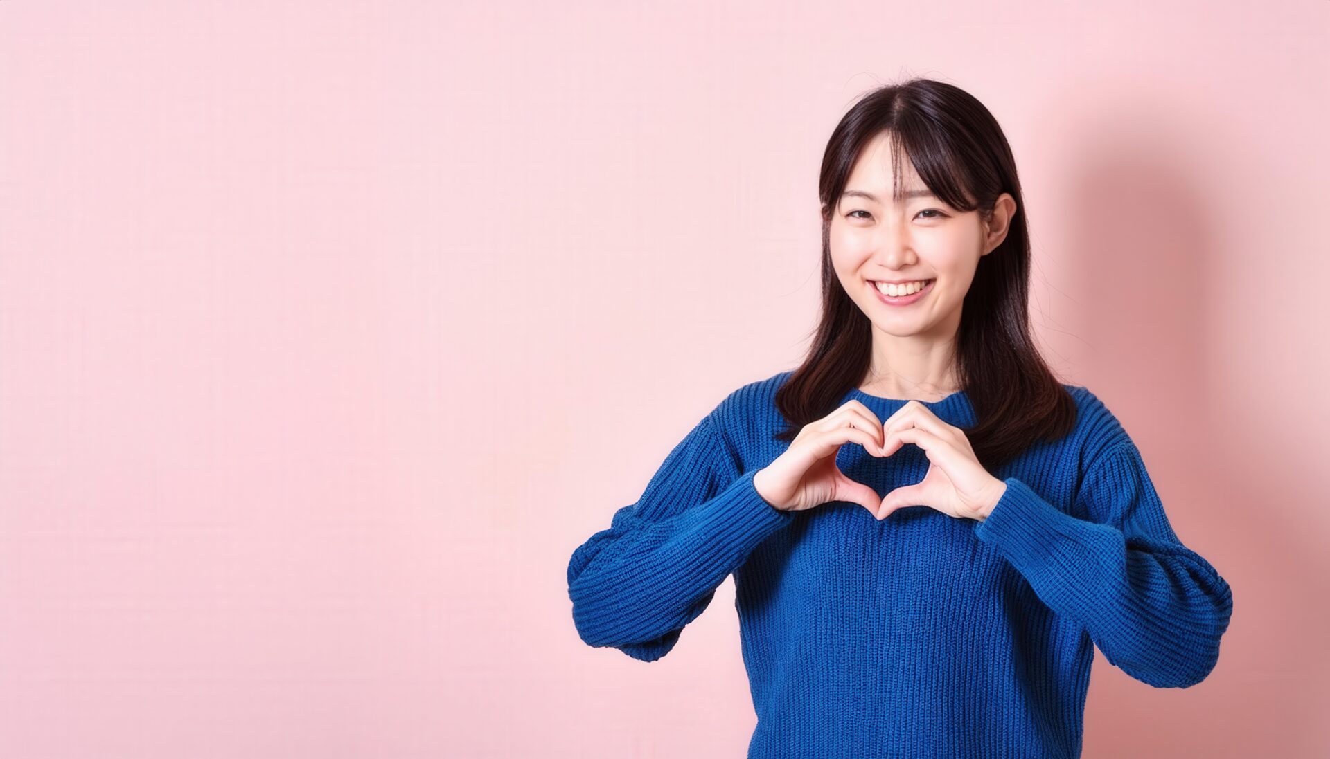 ピンク背景でハートマークを作る若い女性（ブルースウェット）