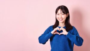 ピンク背景でハートマークを作る若い女性（ブルースウェット）