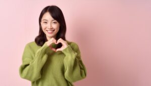 ピンク背景でハートマークを作る若い女性（グリーンスウェット）