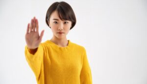 白背景でストップのポーズをする若い女性（イエローニット）