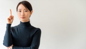白背景で人差し指を立てる若い女性（黒タートルネック）