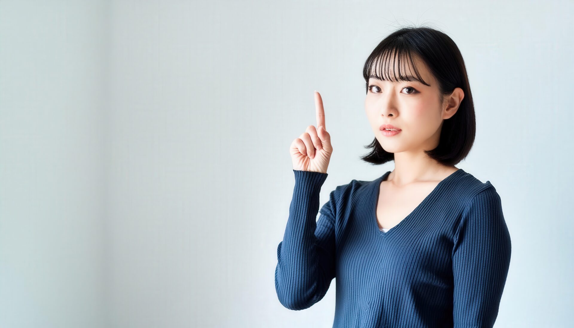 白背景で人差し指を立てる若い女性（ネイビートップス）