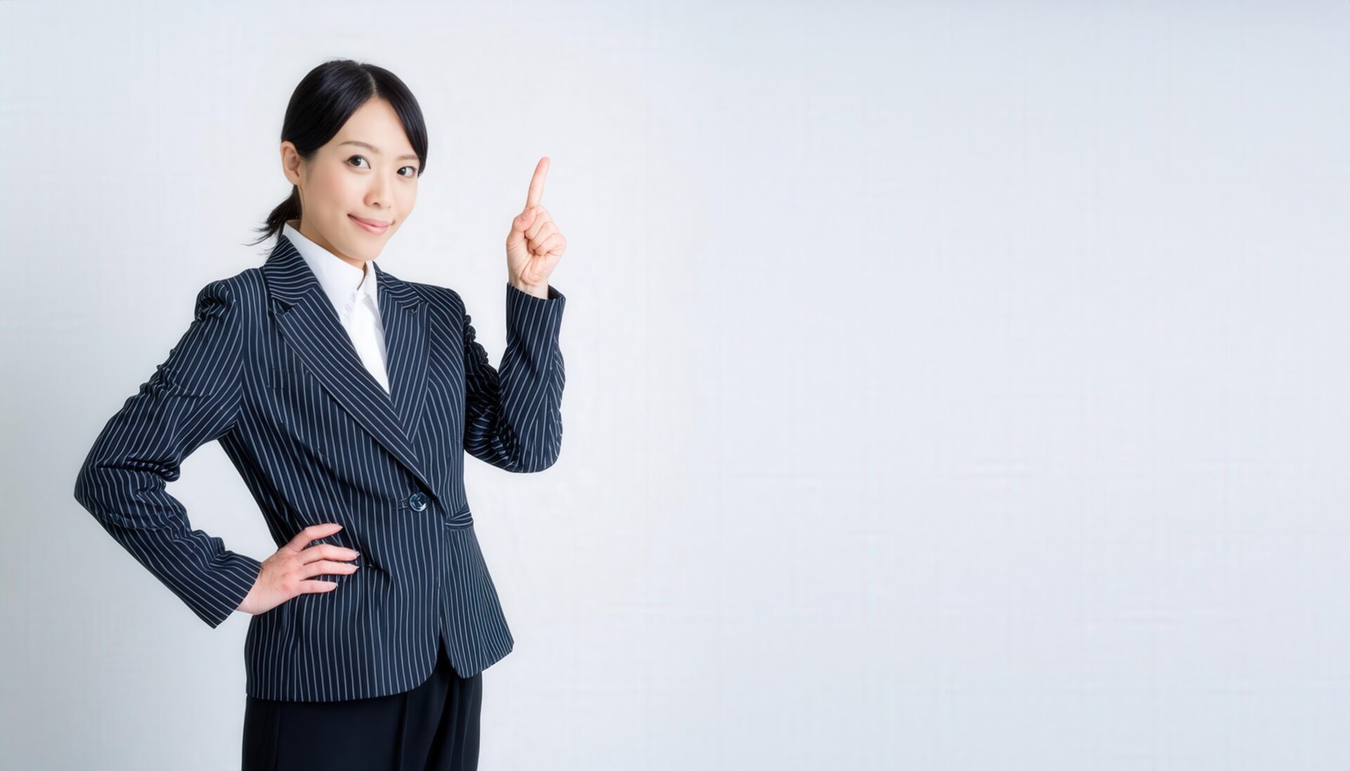 白背景で人差し指を立てる若い女性（ピンストライプスーツ）