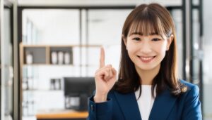 オフィスで人差し指を立てる笑顔の若い女性（ビジネススーツ）