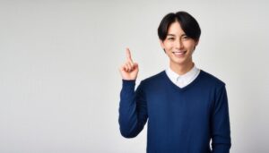 白背景で紺色のセーターを着て人差し指を立てる笑顔の若い男性