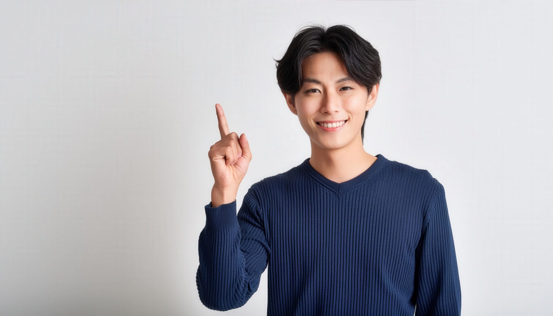 白背景で人差し指を立てる笑顔の若い男性（ネイビーのニット）
