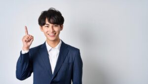 白背景で人差し指を立てる笑顔の若い男性（ストライプスーツ）