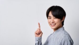 白背景で人差し指を立てる笑顔の若い男性（ストライプシャツ）