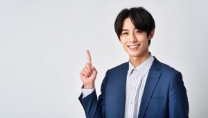 白背景で人差し指を立てる笑顔の若い男性（スーツ×ストライプシャツ）