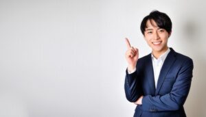 白背景で人差し指を立てる笑顔の若い男性（ビジネススーツ）