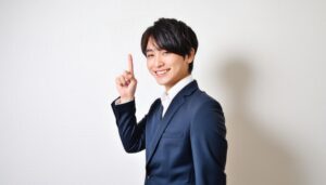 白背景で人差し指を立てる笑顔の若い男性（ネイビースーツ）