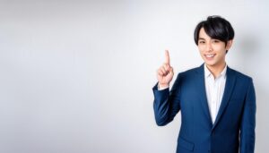 白背景で人差し指を立てる笑顔の若い男性（ブルースーツ）