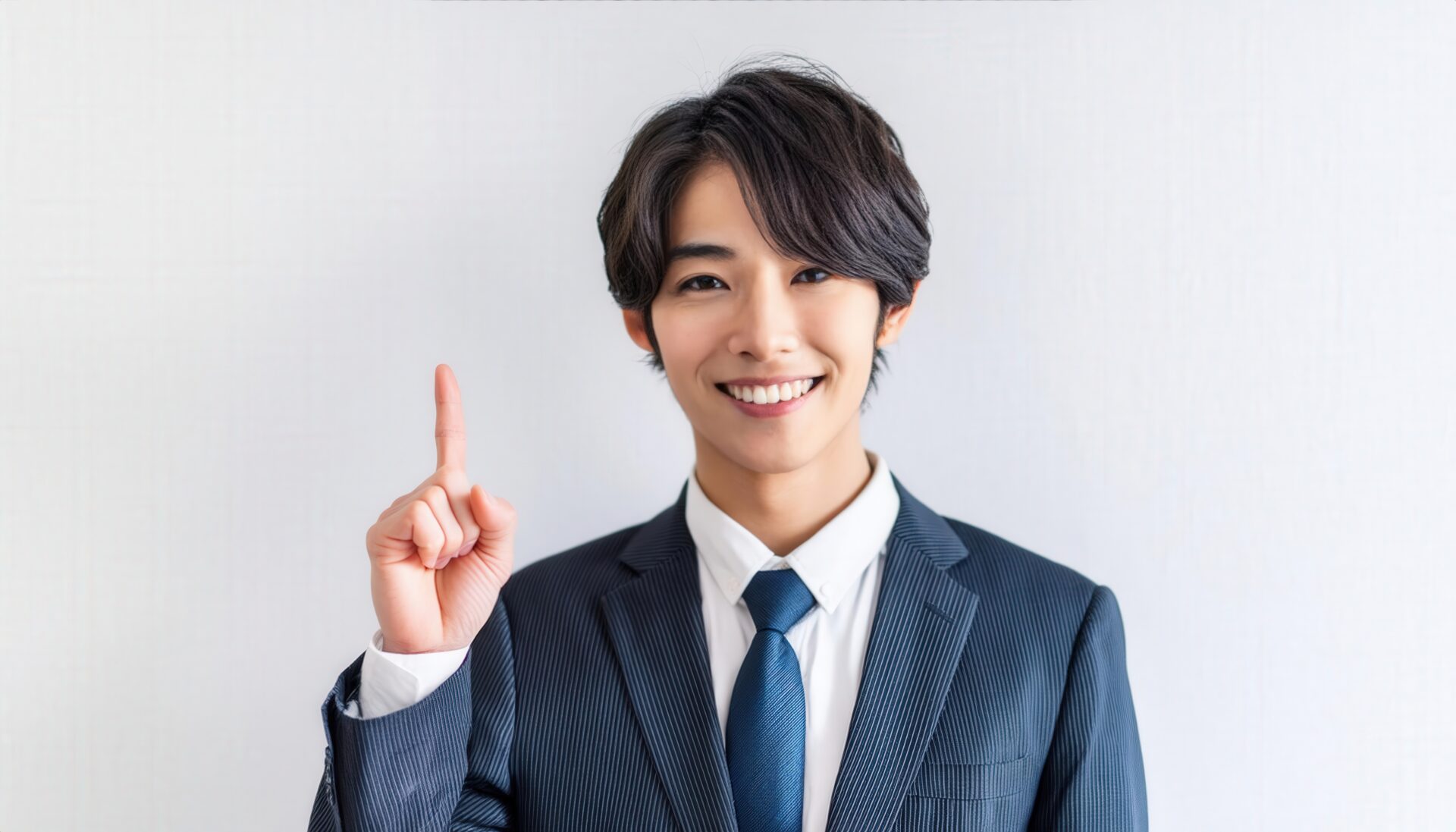 白背景で人差し指を立てる笑顔の若い男性（ビジネスタイ）