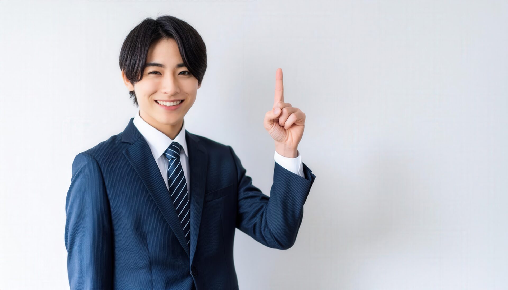 白背景で人差し指を立てる笑顔の若い男性（ネイビースーツ・ストライプタイ）