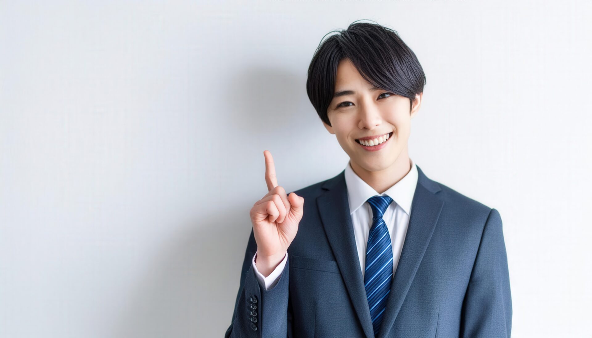 白背景で人差し指を立てる笑顔の若い男性（スーツとブルータイ）