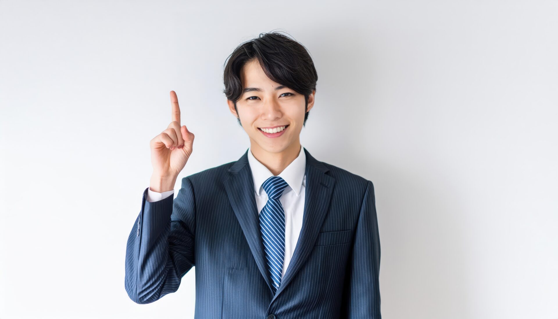 白背景で人差し指を立てる笑顔の若い男性（ダークスーツと青いネクタイ）