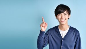 水色背景で人差し指を立てる笑顔の若い男性（ネイビーカーディガン）