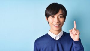 水色背景で人差し指を立てる笑顔の若い男性（紺色のセーター）