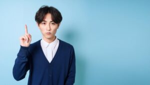 水色背景で人差し指を立てる若い男性（ネイビーカーディガン）