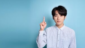 水色背景で人差し指を立てる若い男性（ストライプシャツ）