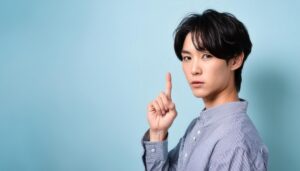 水色背景で人差し指を立てる若い男性（ストライプシャツ）