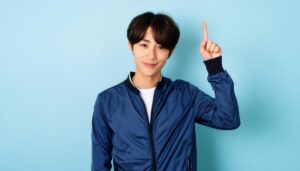 水色背景で人差し指を立てる若い男性（ブルージャンパー）