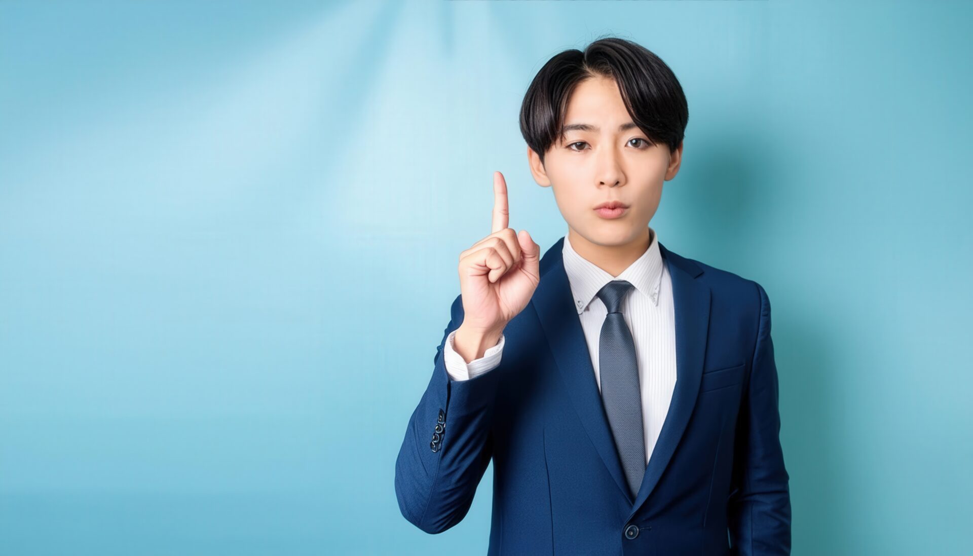 水色背景で人差し指を立てる若い男性（ネイビースーツ）