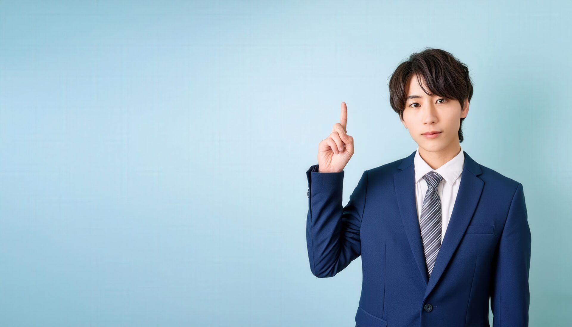水色背景で人差し指を立てる若い男性（ネイビースーツとストライプタイ）