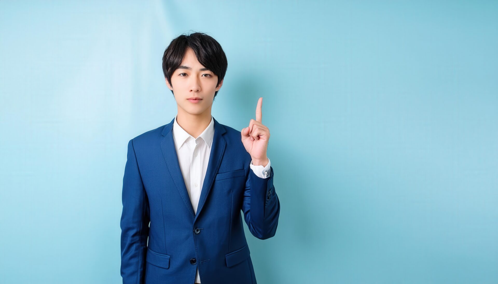 水色背景で人差し指を立てる若い男性（ネイビースーツとワイシャツ）