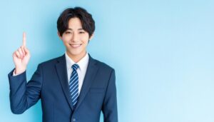 水色背景で人差し指を立てる若い男性（ネイビースーツとブルータイ）