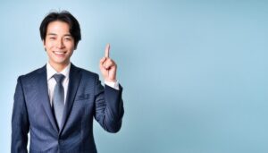 水色背景で人差し指を立てる若い男性（スーツとブルータイ）