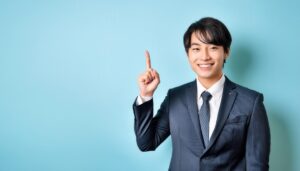 水色背景で人差し指を立てる笑顔の若い男性（スーツ・ダークネクタイ）