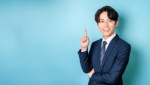 水色背景で人差し指を立てる笑顔の若い男性（ネイビースーツ・ストライプタイ）