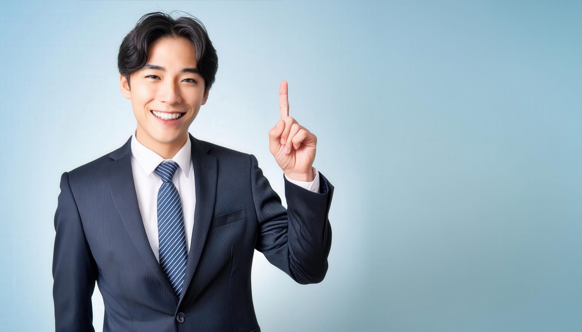 水色背景で人差し指を立てる笑顔の若い男性（ダークスーツ・ストライプタイ）