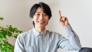 室内で人差し指を立てる笑顔の若い男性（ストライプシャツ）